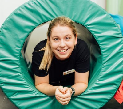 A person with a name badge smiles while lying inside a green circular foam tunnel.