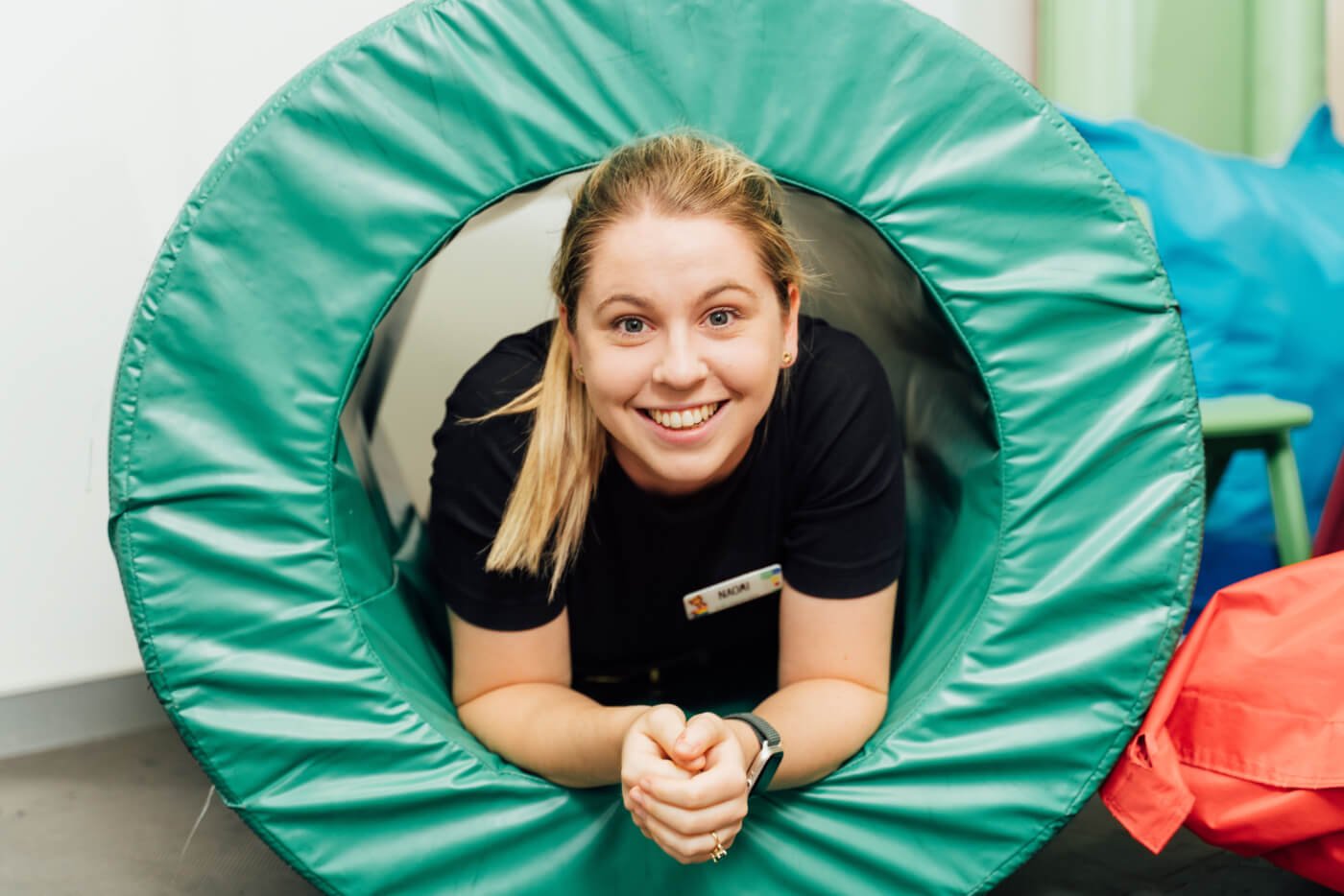 A woman with blonde hair smiles while lying inside a green padded play tunnel.