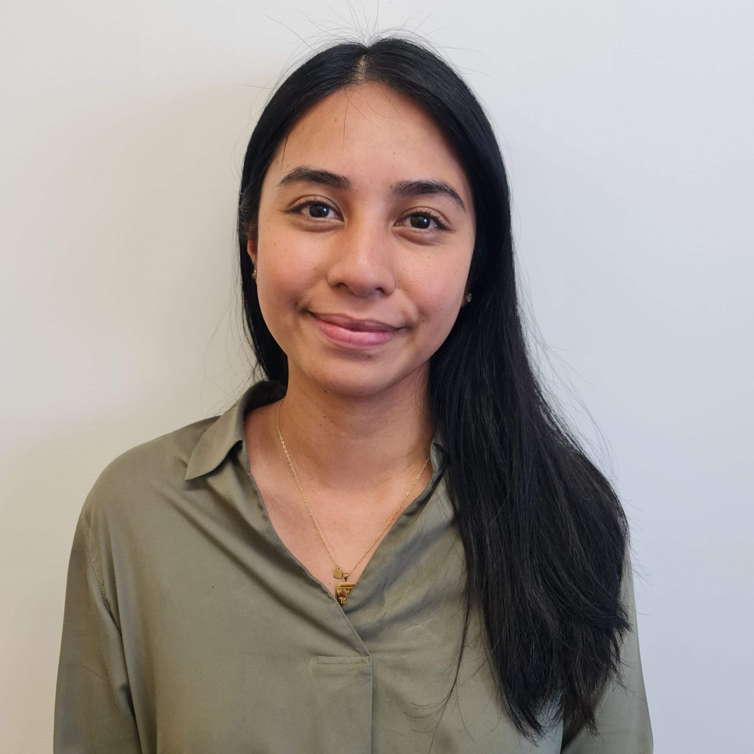 Brooke Szakowski, with long dark hair, wearing a green shirt and a necklace, smiles slightly while standing against a plain white background.