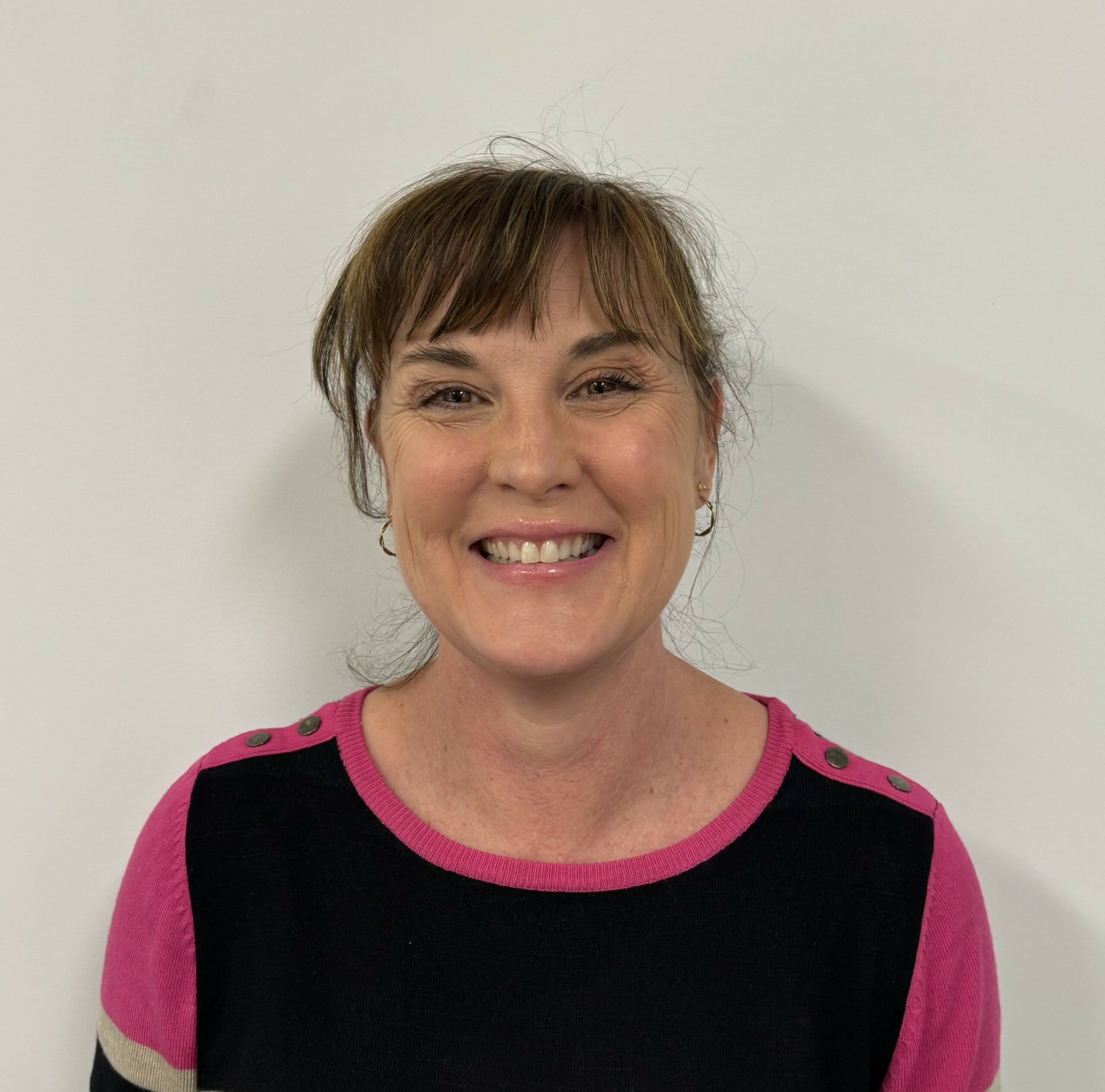 Smiling Fiona Hansen, with light brown hair and bangs, wearing a pink and black shirt, stands against a plain white background.