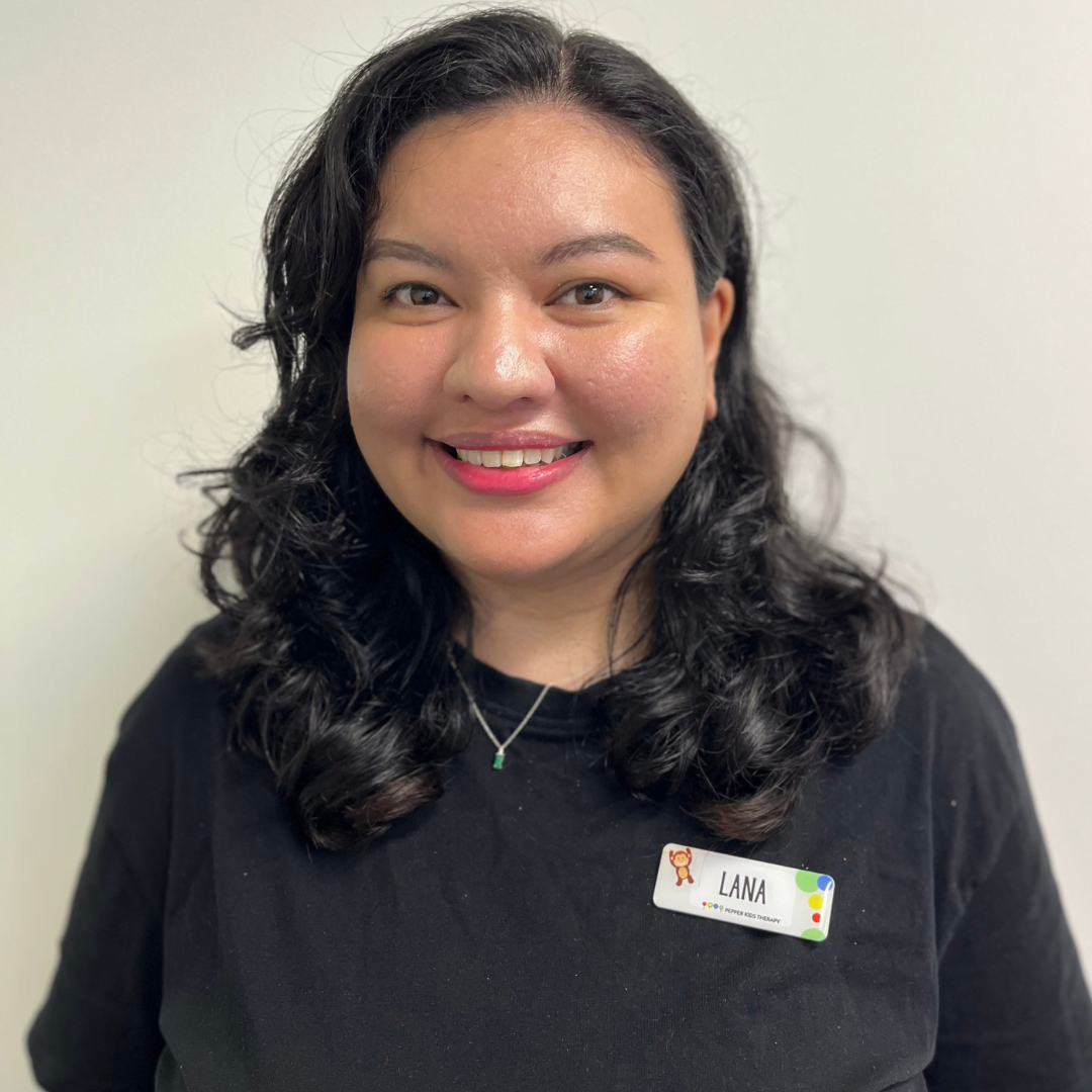 A person with curled dark hair, wearing a black shirt and a name tag that says "Lana London," smiles at the camera against a plain white background.