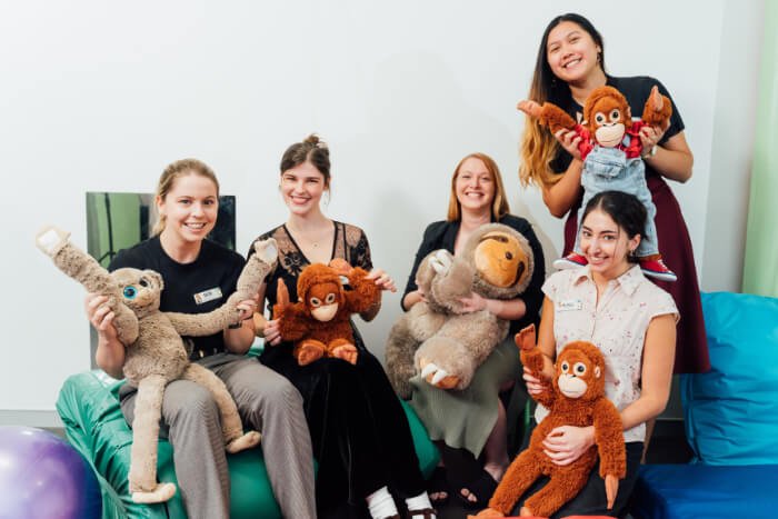 Five women sitting on a couch holding various stuffed animals and smiling at the camera.