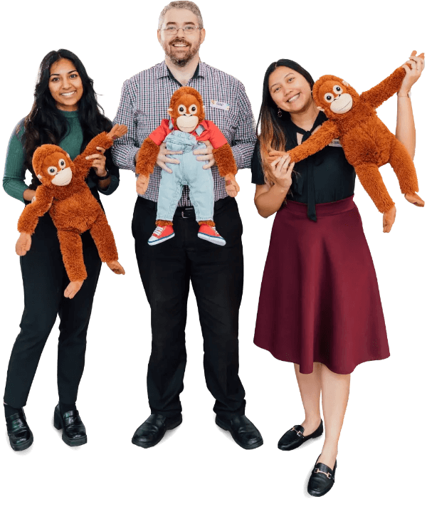 Three people standing, each holding a plush orangutan toy. The man in the middle wears a checked shirt, and the two women on either side are smiling.