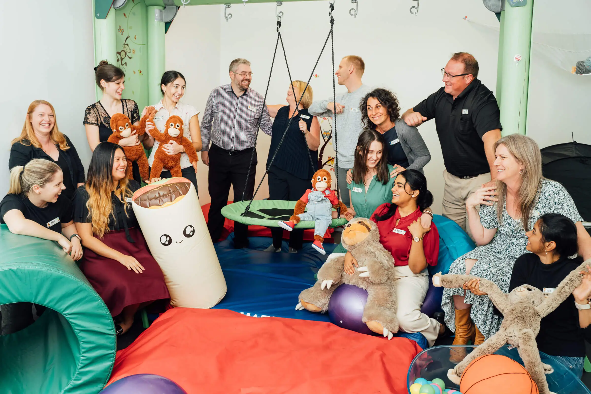 Pepper Kids Therapy team, mostly smiling, in a playroom setting with various toys and playground equipment. Some are holding stuffed animals and interacting in a casual, friendly manner.
