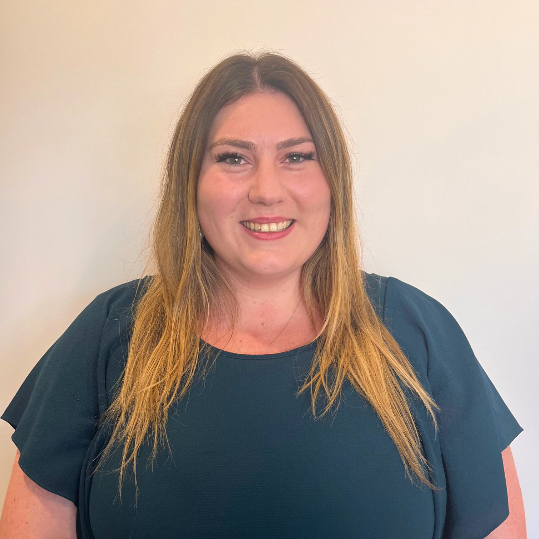 A person with long, straight brown hair wearing a dark green top smiles in front of a plain white background, embodying the elegance and charm associated with Caitlin Bishop-Mowatt.