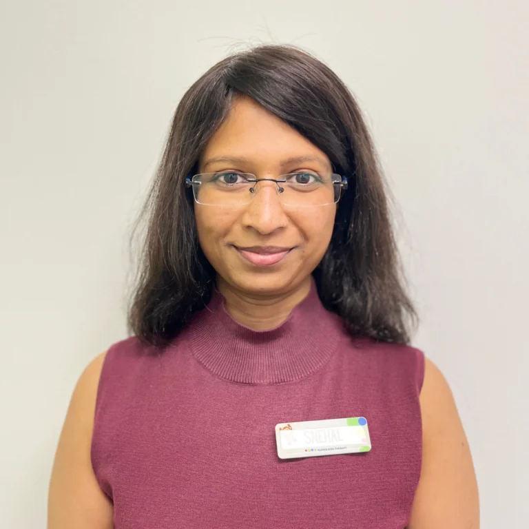 A person with shoulder-length hair and glasses is wearing a maroon sleeveless top and a name badge that reads "SNEHAL VAIDYA" against a plain background.