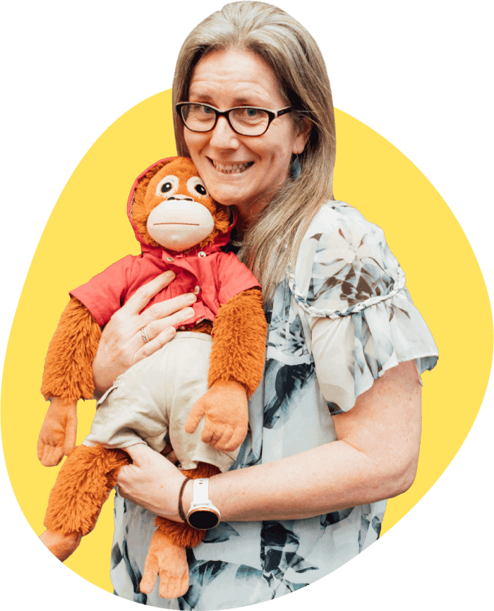 A person wearing glasses and a light-colored floral blouse holds an orange stuffed monkey dressed in a red shirt and beige pants, standing against a yellow background.